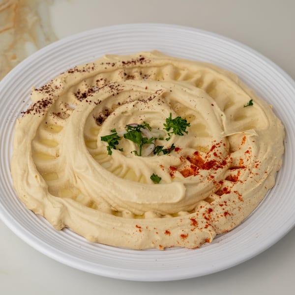 Swirled hummus on a white plate, garnished with olive oil, parsley, red onion, sumac, and paprika.
