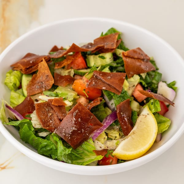 Vibrant Fattoush salad in a white bowl, featuring lettuce, tomatoes, cucumbers, fried pita, and a lemon wedge.